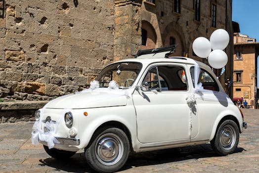 A classic Fiat 500 adorned with wedding decorations and white balloons on a sunny street in Volterra, Italy.